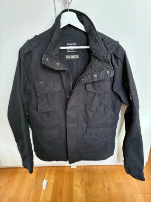 Jacka från Superdry - Snygg svart jacka från Superdry med hög krage, fyra framfickor med lock och dragkedja samt knappar framtill. Jackan har axelklaffar och är tillverkad i ett slitstarkt bomullsmaterial. Perfekt för dig som gillar en stilren och Grisch look. 