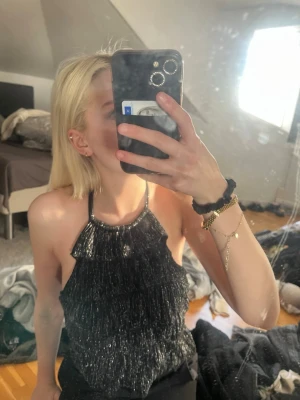 Glittrig svart halterneck topp - Snygg svart glittrig halterneck topp med volangdetaljer och knyt i nacken. Toppen är ärmlös och har en croppad passform som ger en trendig look. Perfekt för dig som vill sticka ut med lite extra glitter.