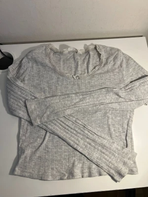 Ljusgrå långärmad topp från H&M - Söt ljusgrå långärmad topp från H&M med diskret ribbad struktur. Rund halsringning dekorerad med spetskant och liten rosett framtill. Perfekt för en enkel och stilren look. Mjuk och bekväm bomullsblandning.