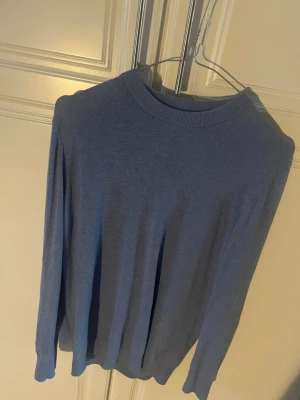 Blå basic tröja med rund hals - En enkel blå tröja med rund halsringning och långa ärmar. Tröjan har ribbade muddar vid hals, ärmslut och nederkant. Perfekt för lager på lager och passar till det mesta. Materialet ser ut att vara mjukt och tunt, troligen bomull eller bomullsblandning.