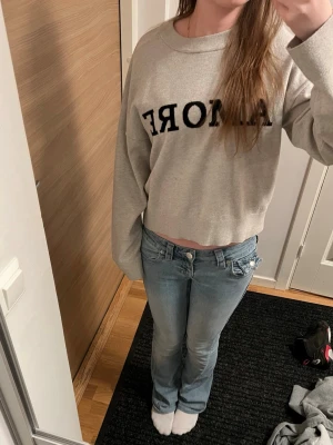 Beige croppad sweatshirt med tryck - Beige croppad tunn stickad sweatshirt med svart texten 'AMORE' framtill. Tröjan har rund halsringning och långa ärmar. Mjuk och bekväm, perfekt för en trendig och avslappnad stil.