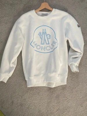 Vit sweatshirt från Moncler - Snygg vit sweatshirt från Moncler med stort blått logotryck på bröstet och klassisk Moncler-patch på ärmen. Tröjan har rund halsringning, ribbade muddar och är tillverkad i mjukt bomullsmaterial. Perfekt för en clean och stilren look.