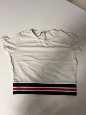 Vit croppad t-shirt med randig kant - Säljer en vit croppad t-shirt med korta ärmar och en bred ribbad kant längst ner i svart, rött och vitt. Perfekt för en avslappnad och trendig look. T-shirten har en enkel rund halsringning och är gjord i mjuk bomull.