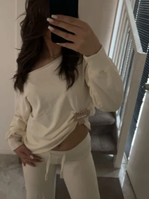 Krämvitt loungewear-set - Mysigt loungewear-set i krämvit färg med långärmad tröja och matchande byxor med dragsko i midjan. Tröjan har avslappnad passform och bred halsringning. Perfekt för chill dagar hemma.