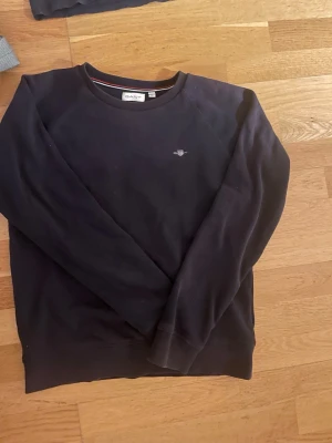 Mörkblå sweatshirt från Gant - Klassisk mörkblå sweatshirt från Gant med rund halsringning och broderad logga på bröstet. Tröjan har ribbade muddar vid ärmslut och nederkant samt en mjuk bomullskänsla. Perfekt för en avslappnad och stilren look.