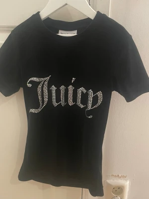 Svart Juicy Couture t-shirt  - Snygg svart t-shirt från Juicy Couture med korta ärmar och stor text 'Juicy' i glittrande strass på bröstet. Klassisk passform och mjukt material som känns skönt mot huden. Perfekt för dig som vill sticka ut med lite extra bling.