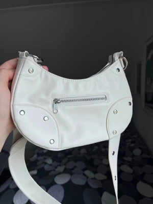 Trendig vit axelväska med nitar – perfekt till sommaren (nyskick) - Snygg vit axelväska med silvriga nitar och dragkedja. Justerbar rem. Rymmer det viktigaste – mobil, plånbok, nycklar. Använd en gång, i mycket fint skick. Mått: ca 24 × 12 cm