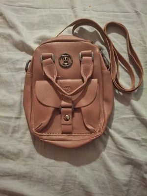 Rosa handväska med axelrem - Söt rosa handväska i konstläder med både handtag och justerbar axelrem. Väskan har en rundad form, ett dekorativt metallmärke med bokstaven B framtill och en ficka med lock och knapp. Perfekt för dig som vill ha en liten och smidig väska. Lite sliten på knappen där fram. Kostar 500kr ny