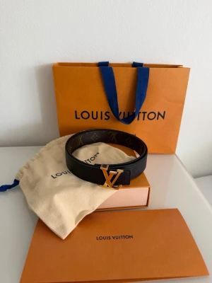 Louis Vuitton svart skärp med LV-spänne - Snyggt svart skärp från Louis Vuitton med klassiskt LV-monogram på insidan och ett guldigt LV-spänne framtill. Skärpet är gjort i exklusivt skinn och har en stilren, tidlös design som lyfter vilken outfit som helst.