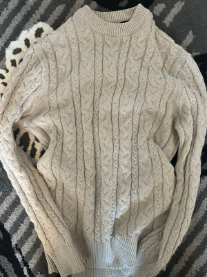 Beige kabelstickad tröja - Snygg beige kabelstickad tröja med hög hals och ribbade muddar. Tröjan har ett klassiskt flätmönster och är perfekt för lager-på-lager-stil. Passar dig som gillar en stilren och mysig look.