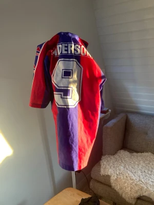 Retro rödblå fotbollströja Anderson - Säljer en retro fotbollströja från Lotto i rött och blått med stora vita siffran 9 och namnet Anderson på ryggen. Tröjan har långa ärmar med Lotto-loggor längs sidorna och klassisk sportig design. Perfekt för dig som gillar vintage sportstil.