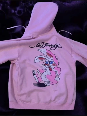 Rosa Ed Hardy hoodie med kaninmotiv - ❗️PRIS KAN DISKUTERAS❗️köpte tröjan för 800kr så vill helst ha runt 400kr. Säljer en rosa hoodie från Ed Hardy med dragkedja och stor huva. På både fram och baksidan finns ett tryck av en tecknad kanin som är detaljerad med diamanter och Ed Hardy-loggan. Perfekt för dig som gillar statement-plagg och Y2K / streetwear-vibe. Säljer tröjan på grund av att jag har bytit stil och därför inte använder tröjan längre. 
