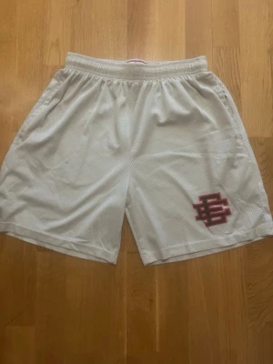 Vita meshshorts med röd logga - Snygga vita meshshorts med röd logga på vänster ben. Shortsen har elastisk midja med snörning och är tillverkade i ett ventilerande polyestermaterial. Perfekta för sport eller chill dagar.