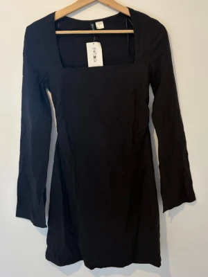 Svart långärmad klänning  - Stilren svart klänning med lång ärm och fyrkantig urringning. Kort modell som ger en modern look. I ett kostym likande material. 