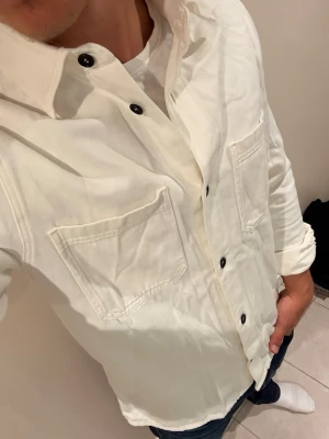 Vit overshirt med svarta knappar - Snygg vit overshirt med raka linjer och två stora bröstfickor. Skjortan har svarta knappar framtill och vid ärmsluten, samt klassisk krage. Perfekt att bära öppen över en t-shirt för en avslappnad look.