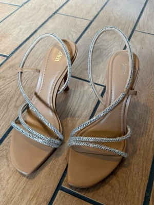 Beige klackskor med strass från Zara - Snygga beige klackar från Zara med smala remmar dekorerade med glittrande strass. Klackarna har fyrkantig tå och hög klack, vilket ger en trendig och elegant look. Perfekta för att lyfta din outfit med lite extra bling. Helt oanvända. 