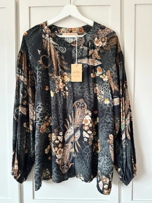 Mönstrad viskosblus från Marta du Chateau L/XL - Snygg svart blus med paisley- och blommönster i brunt, beige och orange från Marta du Chateau. Blusen har rund halsringning, knappar framtill och långa, lösa ärmar. Materialet är mjukt och följsamt, perfekt för en trendig look. 100% viskos. 