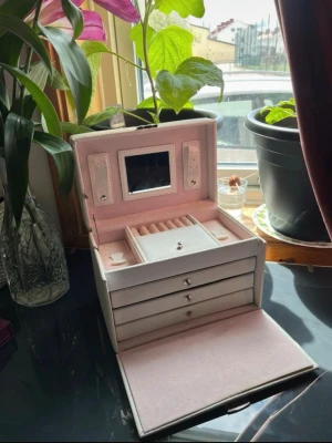 Ljusrosa smyckeskrin med spegel - Snyggt smyckeskrin i ljusrosa konstläder med flera lådor och fack, perfekt för att organisera ringar, örhängen och halsband. Insidan är klädd i mjuk sammet och locket har en liten spegel. Praktisk och stilren förvaring för dina accessoarer.