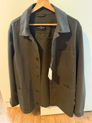 Mörkgrön overshirt från John Henric - Stilren mörkgrön overshirt från John Henric med klassisk krage och tre stora utanpåfickor. Overshirten har knäppning framtill och är tillverkad i ett mjukt, slitstarkt bomullstyg. Perfekt lager-på-lager-plagg för en avslappnad look.