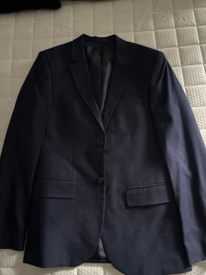 Mörkblå kavaj från h&m - Stilren mörkblå kavaj från Filippa K med två knappar framtill och klassisk krage. Kavajen har två fickor med lock och fyra knappar vid ärmslutet. Materialet är slätt och ger en dressad look, perfekt för en snygg och modern stil.