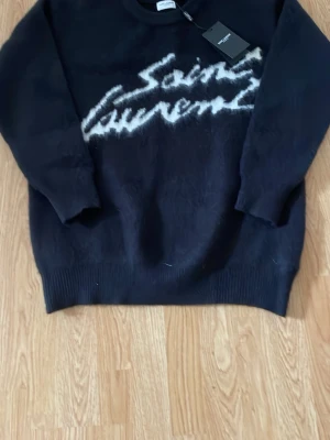  Saint Laurent stickad tröja - Svart stickad tröja från Saint Laurent med vit, fluffig logotyp framtill. Tröjan har rund halsringning och ribbstickade muddar. Perfekt för dig som vill ha en lyxig och trendig look med streetkänsla.