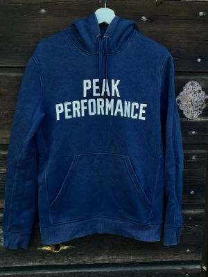 Blå Peak Performance hoodie - Mörkblå hoodie från Peak Performance med vit logga tryckt på bröstet. Klassisk modell med huva och dragsko samt stor magficka framtill. Tillverkad i mjukt bomullsmaterial och har ribbade muddar vid ärmslut och nederkant.