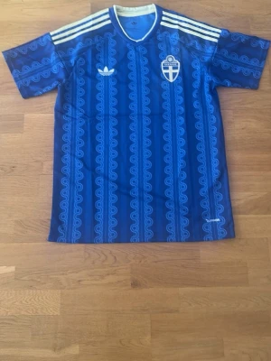 Blå Sverige fotbollströja Adidas - Säljer en blå fotbollströja från Adidas med Sveriges landslagsemblem på bröstet. Tröjan har vita ränder på axlarna, mönstrad design i olika blå nyanser och klassisk Adidas-logga. Perfekt för fotboll eller träning.