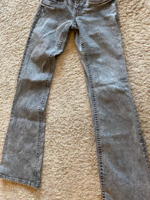 Grå raka jeans med klassisk passform - Säljer ett par grå jeans med utsvängda ben och klassisk femficksdesign. Jeansen har normal midja och är tillverkade i denim. Perfekta för en avslappnad och stilren look.