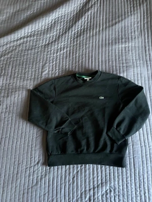 Svart sweatshirt från Lacoste  - En stilren svart sweatshirt från Lacoste med klassisk rund halsringning och det ikoniska krokodilmärket broderat på bröstet. Tröjan har långa ärmar, ribbade muddar och är gjord i mjukt bomullsmaterial. Perfekt för en avslappnad och trendig look.