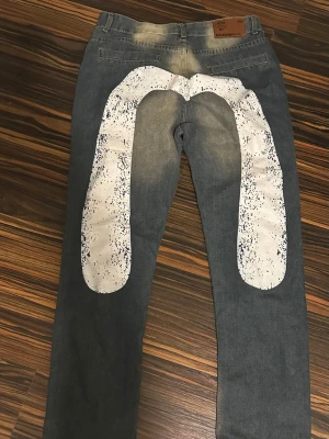 Grå jeans med vit print och slitningar - Unika grå jeans med stor vit print på baksidan och slitna detaljer framtill. Jeansen har en tvättad look och raka ben. Materialet är klassisk denim och de har fem fickor samt knappgylf. Perfekta för dig som vill sticka ut med din stil.