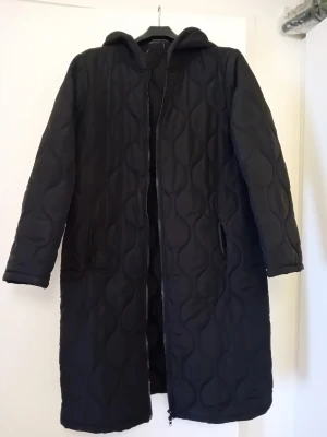 Svart quiltad lång kappa med huva - Snygg svart quiltad kappa med huva och dragkedja framtill. Lång modell med två fickor på sidorna och vågigt mönster i tyget. Perfekt för kyliga dagar och ger en stilren look.