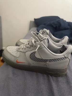Nike Air Force 1 grå sneakers - Grå Nike Air Force 1 sneakers med vit ovandel, grå detaljer och en orange mini-swoosh framtill. Swoosh-loggan på sidan är i grått meshmaterial för en sportig look. Klassisk låg modell med snörning och perforerad tå för ventilation.