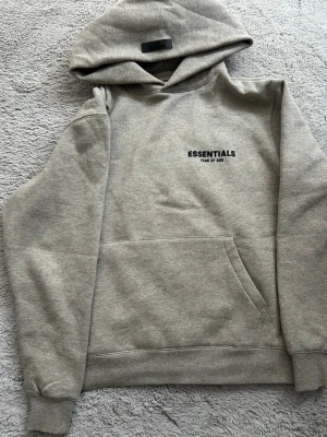 Grå Essentials Fear of God hoodie - Snygg grå hoodie från Essentials Fear of God med svart logga på bröstet. Klassisk modell med huva och stor magficka. Perfekt för dig som gillar streetwear och vill ha en stilren look. Mjuk och skön bomullsblandning.