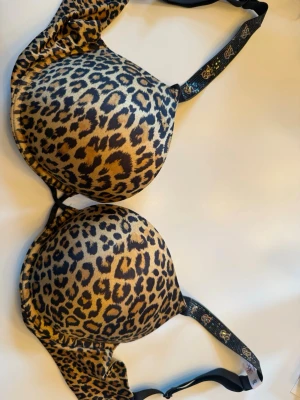 Leopardmönstrad push-up bh från Victoria's Secret - Snygg push-up bh från Victoria's Secret med leopardmönster i brunt och svart. Axelbanden är dekorerade med glittrande detaljer och VS-loggor. Kuporna är vadderade för extra lyft. Helt oanvänd!