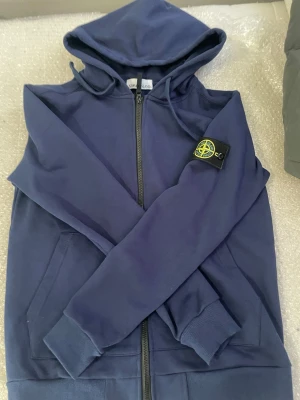 Blå hoodie från Stone Island - Snygg blå hoodie från Stone Island med dragkedja och klassisk logotyp-patch på ärmen. Hoodien har huva med dragsko, ribbade muddar och fickor framtill. Perfekt för en avslappnad och trendig stil.