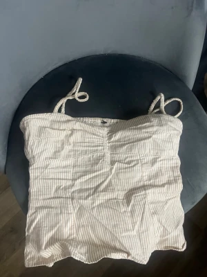 Beige och vit randig linnetopp - Söt linnetopp med tunna axelband som knyts, i beige och vitt randigt mönster. Toppen har smockad rygg och volangdetalj nedtill. Perfekt för varma dagar och enkel att styla till jeans eller kjol.