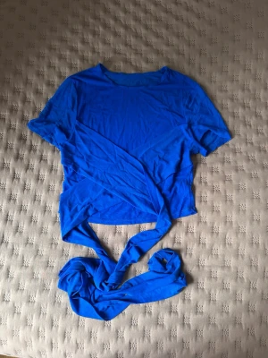 Blå mesh omlottopp med knytband - Snygg blå mesh topp med korta ärmar och långa knytband i midjan som ger en omlott-effekt. Tunt och genomskinligt material som känns lätt och luftigt. Märket är tyvärr okänd på grund av att jag klippt av lappen där även material specifikationen stod.