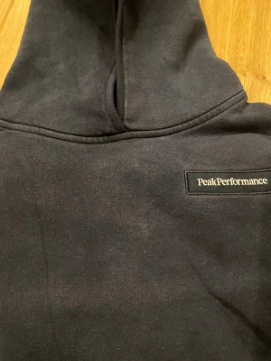 Mörkblå hoodie från Peak Performance - Mörkblå hoodie från Peak Performance med klassisk logga på bröstet. Hoodien har en stor huva och en magficka framtill. Tillverkad i mjukt bomullsmaterial och har ribbade muddar vid ärmslut och nederkant. Perfekt för en avslappnad och stilren look.