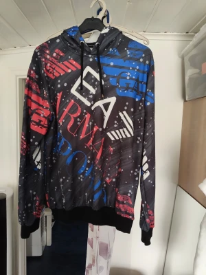 Färgglad hoodie från Armani Exchange - Snygg hoodie från Armani Exchange med svart bas och stora tryck i rött, blått och vitt. Huva med dragsko och ribbade muddar. Perfekt för dig som gillar streetwear och vill sticka ut med en unik design.
