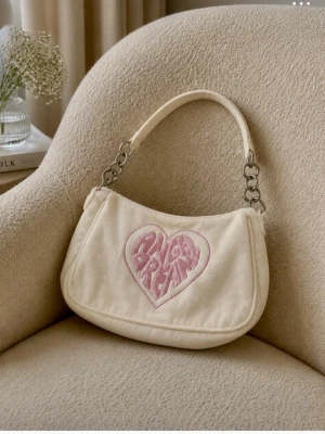 Vit axelväska med rosa text - Säljer en söt vit axelväska med texten 'Angel Dream' i rosa på framsidan. Väskan har en kedjedetalj på axelremmen och är perfekt för att ge en touch av stil till din outfit. Den är tillverkad i ett mjukt material och har en dragkedja för säker förvaring.