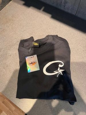 Svart t-shirt från Adventure S - Svart t-shirt från Adventure med en stor vit tryckt logga på bröstet som föreställer ett C och en stjärna. Klassisk rund halsringning och mjukt material som känns skönt mot huden. Perfekt för en chill och stilren look.