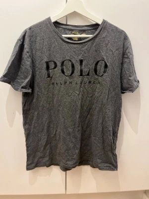 Grå t-shirt från Polo Ralph Lauren - Snygg grå t-shirt från Polo Ralph Lauren med svart tryck på bröstet. Klassisk rund halsringning och korta ärmar. Perfekt för en avslappnad och stilren look.