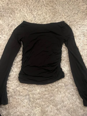 Svart off shoulder topp med långa ärmar - Snygg svart off shoulder topp med långa ärmar och rynkade sidor. Toppens material är stretchigt och sitter tajt mot kroppen, perfekt för en trendig look. Passar dig som gillar stilrena och enkla plagg med en twist.