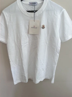 Vit Moncler t-shirt med logga - Snygg vit t-shirt från Moncler med klassisk rund hals och broderad logga på bröstet. Tillverkad i mjuk bomull och har en stilren, enkel design som passar till allt. Perfekt för dig som gillar exklusiva och tidlösa plagg.