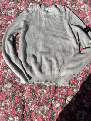 Ljusgrå stickad tröja Stone Island - Snygg ljusgrå stickad tröja från Stone Island med rund halsringning och ribbade muddar. Klassisk och stilren design, perfekt att matcha med jeans eller chinos. Tröjan har lång ärm och är tillverkad i ett mjukt, ribbstickat material. Om man fotar lappen är qr Koden redo att komma in på sidan 