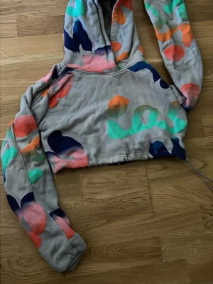 Färgglad cropped hoodie från H&M Studio - Säljer en grå cropped hoodie från H&M Studio A/W 2021 med färgglada abstrakta mönster i orange, grönt, rosa och blått. Hoodien har huva och långa ärmar, perfekt för en trendig och avslappnad stil.