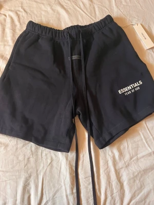 Svarta shorts Essentials Fear of God - Svarta shorts från Essentials Fear of God med vit logga på vänster ben. Modellen har hög midja, långa snören i midjan och är tillverkad i mjuk bomull. Perfekta för en avslappnad och trendig stil.