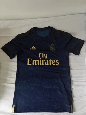Real Madrid bortatröja Adidas blå - Mörkblå Real Madrid bortatröja från Adidas med guldfärgade detaljer och Fly Emirates-tryck på bröstet. Tröjan har korta ärmar, rund hals och klubbmärke på vänster sida. Tillverkad i lätt och ventilerande material, perfekt för fotbollsträning.