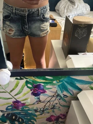 Blå slitna jeansshorts med hög midja - Snygga blå jeansshorts med hög midja och slitna detaljer framtill. Shortsen har klassisk femficksdesign och råa kanter nedtill för en trendig look. Perfekta för varma sommardagar och enkel att matcha med olika toppar. 