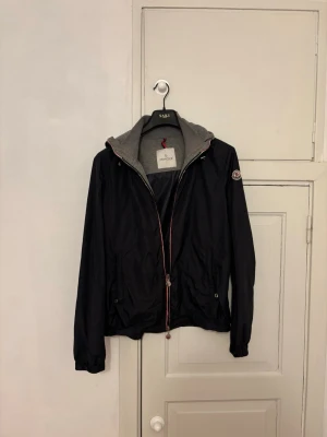 Svart vindjacka från Moncler - Snygg svart vindjacka från Moncler med grå huva och dragkedja med rödvit detalj. Jackan har två fickor med knappar och Moncler-logga på ärmen. Perfekt för blåsiga dagar och enkel att matcha med olika outfits. Äkta förstås. Hör av dig vid frågor eller fler bilder!!!
